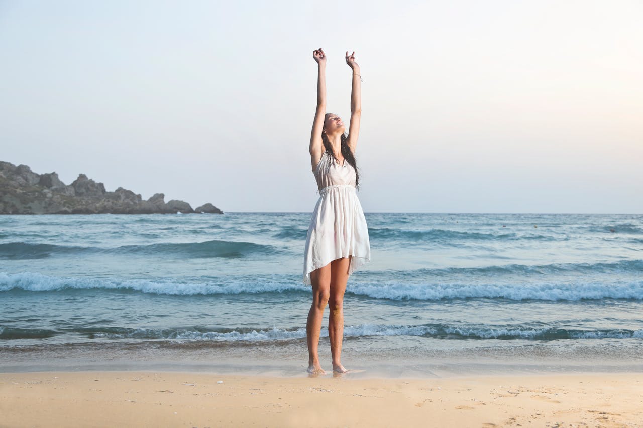 A woman in a white dress stretches joyfully on the sandy beach of Mgarr, Malta, during a serene summer day.